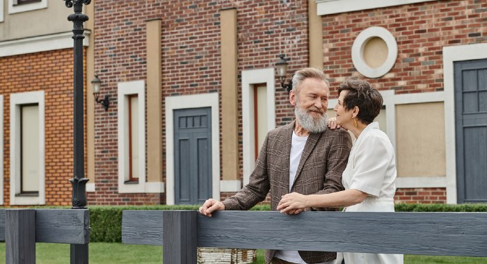 older couple standing at a fence in front of a house smiling at each other